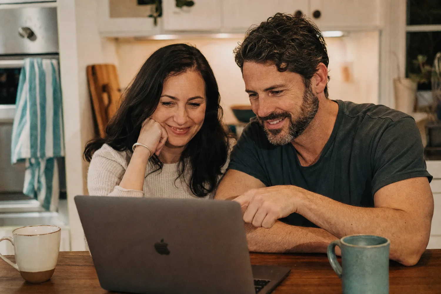 A brother and sister sit at a kitchen table looking at a laptop together, smiling.